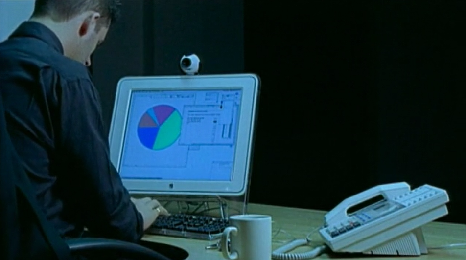 A man sitting at an office desk, typing on a computer which shows a pie chart on the screen.  There is a landline telephone on the desk to to his right.