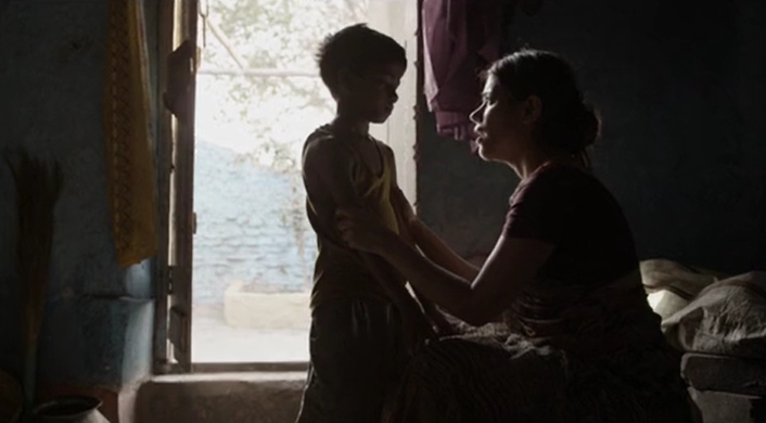 Lit by an open doorway, a sitting woman gently holds a standing small boy by his arms as she speaks to him.