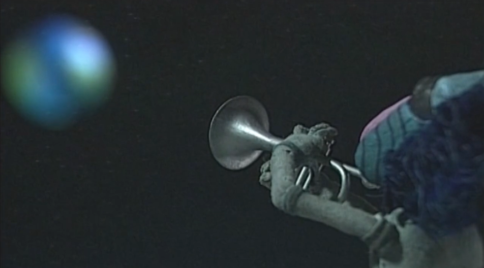 A Claymation baboon plays a silver trumpet, with a blurred view of the Earth in a dark sky in the background.