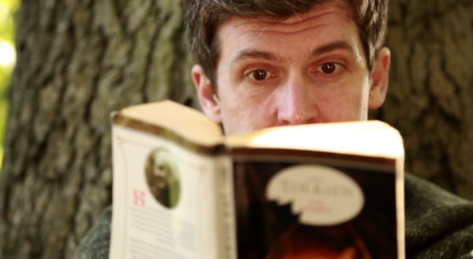 Close-up of a man reading a book, sitting against a tree trunk.