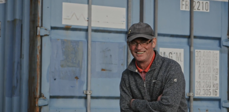 A middle-aged man wearing a muted blue cardigan and baseball cap stands laughing in front of a large metal storage unit.