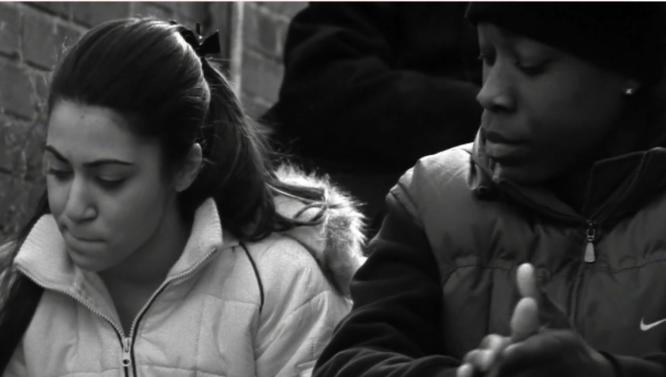 A teenage girl and boy in puffa jackets sit together, looking down, in a black and white still