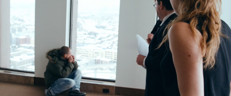 A man crouches on the ground next to large skyscraper windows, watched by three office workers.